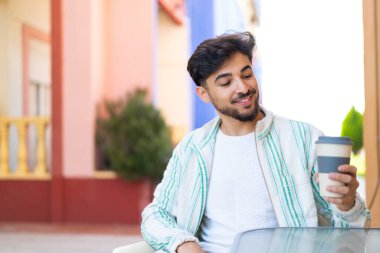 Yakışıklı bir Arap, açık havada elinde kahveyle neşeli bir ifade takınıyor.