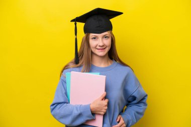 Sarı arka planda izole edilmiş genç bir bayan kolları kalçasında ve gülümsüyor.