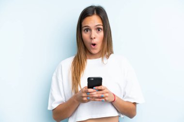 Mavi arka planda izole edilmiş genç beyaz kadın kameraya bakarken şaşırmış bir ifadeyle cep telefonunu kullanıyor.