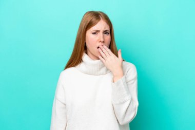 Genç kızıl saçlı kadın mavi arka planda izole edilmiş esniyor ve ağzını eliyle açık tutuyor.