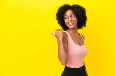Sarı arka planda izole edilmiş genç Afrikalı Amerikalı kadın bir ürünü sunmak için kenarı işaret ediyor.