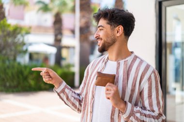Dışarıda cüzdan tutan yakışıklı bir Arap, bir ürünü sunmak için yan tarafı işaret ediyor.
