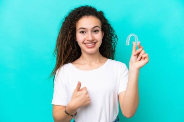 Mavi arka planda görünmez diş telleri tutan genç bir Arap kadın. Başparmakları havada. Çünkü iyi bir şey oldu.