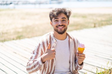 Açık havada kornet dondurması olan yakışıklı bir Arap. Başparmakları havada. Çünkü iyi bir şey oldu.