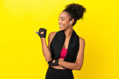 Sarı arka planda izole edilmiş genç Afrikalı sporcu kadın bir ürün sunmak için kenarı işaret ediyor.