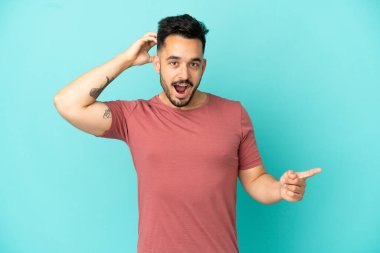 Kafkasyalı genç adam mavi arka planda izole edildi. Şaşırdı ve işaret etti.