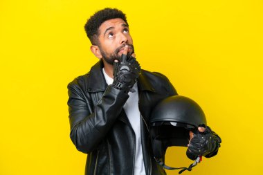 Motosiklet kasklı genç Brezilyalı adam sarı arka planda izole edilmiş, yukarı bakarken şüpheleri var.