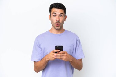 İzole edilmiş beyaz arka plan üzerinde genç yakışıklı bir adam kameraya bakarken şaşırmış bir ifadeyle cep telefonuna bakıyor.