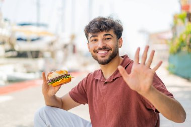 Genç Arap yakışıklı bir adam elinde hamburgerle elini sallayarak mutlu bir ifadeyle selam veriyor.