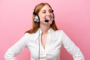 Telemarketer kızıl saçlı kadın pembe arka planda izole edilmiş bir kulaklıkla çalışıyor. Kolları kalçasında ve gülümsüyor.