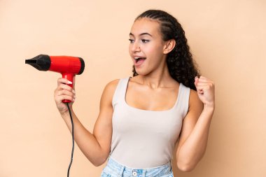 Elinde saç kurutma makinesi olan genç bir kadın zafer kutlamaları için bej arka planda izole edilmiş.