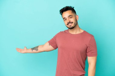 Mavi arka planda izole edilmiş genç beyaz adam, davet edilmek için ellerini yana uzatıyor.