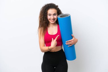 Genç Arap spor kadını yoga derslerine giderken beyaz arka planda izole edilmiş bir minder tutuyor ve bir ürünü sunmak için yan tarafı işaret ediyor.