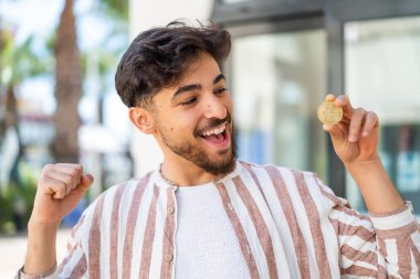 Yakışıklı bir Arap elinde Bitcoin ile dışarıda zaferi kutluyor.
