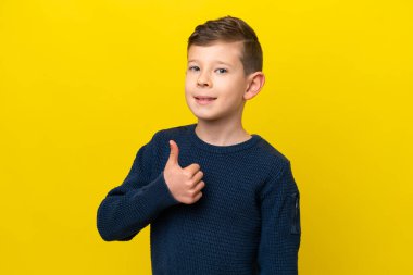 Küçük beyaz çocuk sarı arka planda izole edilmiş bir şekilde baş parmak hareketi yapıyor.