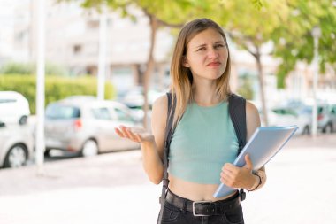 Açık havada genç sarışın bir bayan omuzlarını kaldırırken şüphe jesti yapıyor.