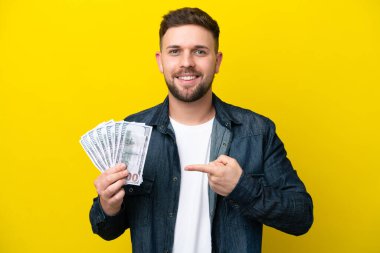 Young caucasian man taking a lot of money  isolated on yellow background pointing to the side to present a product