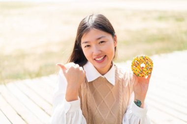 Genç Çinli bir kadın açık havada donut tutuyor ve bir ürün sunmak için kenarı işaret ediyor.