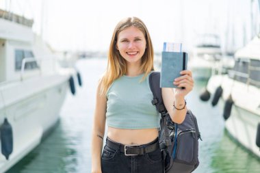 Genç sarışın kadın elinde pasaportuyla dışarıda mutlu bir ifadeyle bekliyor.