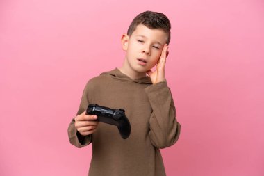 Pembe arka planda izole edilmiş ve baş ağrısı olan bir oyun kontrolörüyle oynayan beyaz bir çocuk.