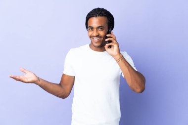 Örgülü genç Afrikalı Amerikalı adam mor arka planda izole edilmiş cep telefonuyla biriyle sohbet ediyor.