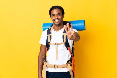 Sarı arka planda büyük bir sırt çantalı genç dağcı adam mutlu ve parmakları olan üçe kadar sayıyor.