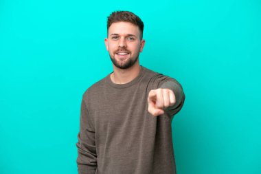 Mavi arka planda izole edilmiş genç beyaz adam mutlu bir ifadeyle ön tarafı işaret ediyor.