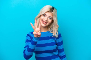Mavi arka planda izole edilmiş genç Rus kadın mutlu ve parmaklarıyla üçe kadar sayıyor.