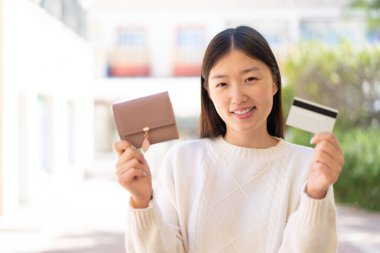 Dışarıdaki güzel Çinli kadın elinde cüzdan ve mutlu bir ifadeyle kredi kartı tutuyor.