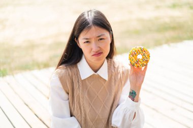 Genç Çinli bir kadın dışarıda üzgün bir ifadeyle donut tutuyor.