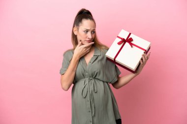Pembe arka planda izole edilmiş genç bir kadın hamile ve elinde şüpheleri varken bir hediye tutuyor.