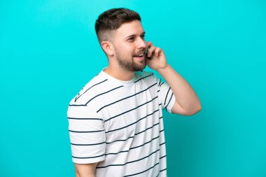 Genç beyaz adam mavi arka planda izole edilmiş cep telefonuyla sohbet ediyor.