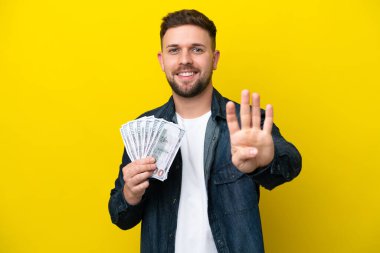Young caucasian man taking a lot of money  isolated on yellow background happy and counting four with fingers
