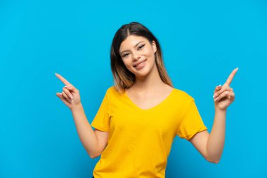 Mavi arka planda izole edilmiş genç Rus kız parmak uçlarını işaret ediyor ve mutlu.