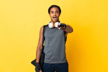 Sarı arka planda izole edilmiş çantalı Afrikalı Amerikalı genç adam şaşırmış ve ön tarafı işaret ediyor.