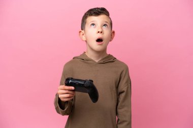 Pembe arka planda izole edilmiş bir bilgisayar oyunu kontrolörüyle oynayan küçük beyaz çocuk yukarı bakıyor ve şaşırmış bir ifadeyle