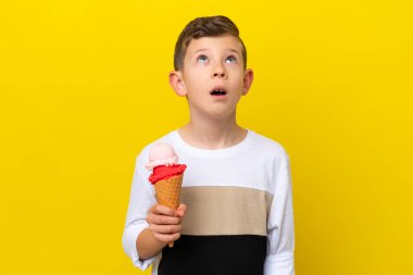 Kornet dondurmalı küçük beyaz çocuk sarı arka planda izole edilmiş yukarı bakıyor ve şaşırmış bir ifadeyle