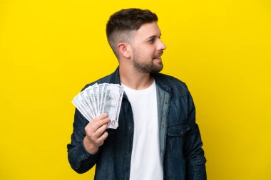 Kafkasyalı genç adam, sarı arka planda izole edilmiş bir şekilde bir sürü para alıyor. Yana bakıyor ve gülümsüyor.
