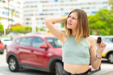 Açık havada arabanın anahtarlarını tutan genç sarışın bir kadın. Şüpheleri var ve kafası karışmış bir yüz ifadesi var.