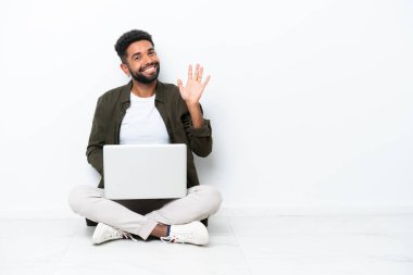 Yerde dizüstü bilgisayarı olan genç Brezilyalı bir adam, beyaz tenli, mutlu bir ifadeyle selam veriyor.