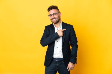 İzole edilmiş sarı arka planda iş adamı bir ürünü sunmak için kenarı işaret ediyor.