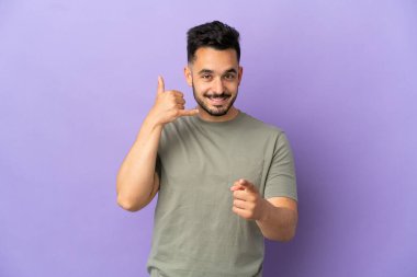 Mor arka planda izole edilmiş genç beyaz bir adam telefon jesti yapıyor ve ön tarafı işaret ediyor.