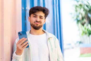 Yakışıklı bir Arap açık havada üzgün bir ifadeyle cep telefonu kullanıyor.