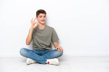 Genç Rus adam, beyaz arka planda izole bir şekilde yerde oturuyor. Parmaklarıyla işaret veriyor.