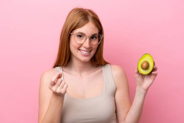 Genç kızıl saçlı kadın pembe arka planda izole edilmiş bir avokado tutuyor ve para hareketi yapıyor.