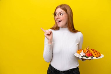 Sarı arka planda izole edilmiş waffle tutan genç kızıl saçlı kadın çözümü bulmak için parmağını kaldırıyor.