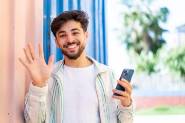 Yakışıklı bir Arap açık havada cep telefonu kullanıyor ve mutlu bir ifadeyle selamlıyor.