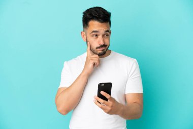 Kafkasyalı genç adam mavi arka planda cep telefonu kullanıyor ve düşünüyor.