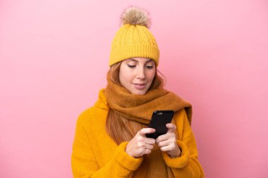 Kışlık ceket giyen genç kızıl saçlı kadın pembe arka planda izole edilmiş cep telefonuyla bir mesaj gönderiyor.
