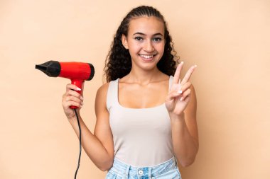 Elinde bej arka planda izole edilmiş saç kurutma makinesi tutan genç bir kadın gülümsüyor ve zafer işareti gösteriyor.